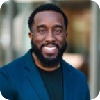 Smiling man with beard wearing a blue blazer and black shirt outdoors with blurred background.