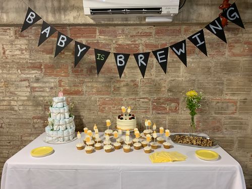 Baby shower table with cupcakes, a diaper cake, cookies, and a "Baby is Brewing" banner against a rustic brick wall.