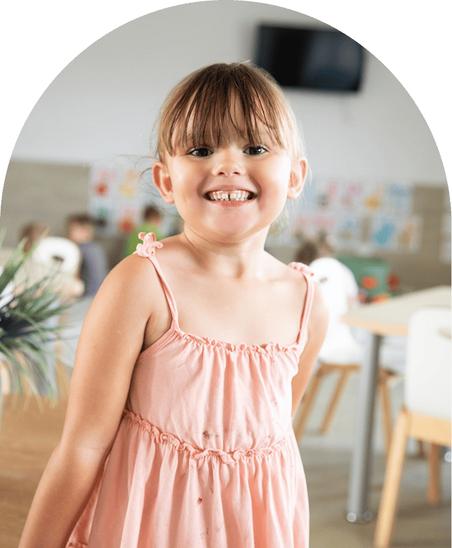 Smiling young girl wearing a pink dress standing indoors with blurred colorful artwork and furniture in the background.