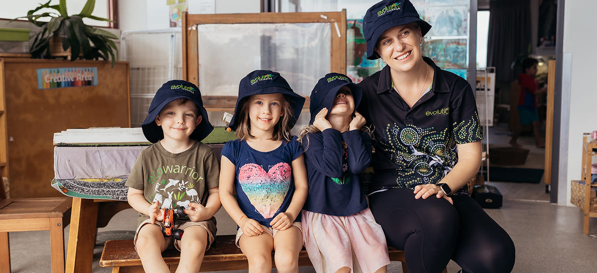 Three children and one adult wearing matching black hats and sitting on a bench inside a classroom.