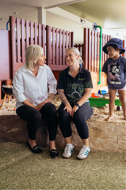 Two women sitting and smiling while a child stands nearby in a play area with sand and wooden fencing.