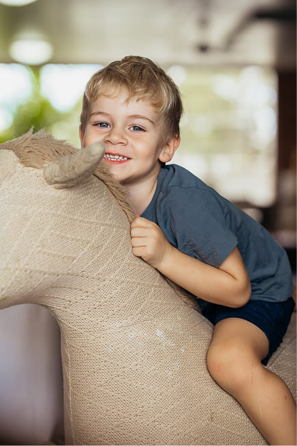 Smiling young boy with blonde hair wearing a blue shirt, hugging and sitting on a large knitted stuffed animal resembling a horse.