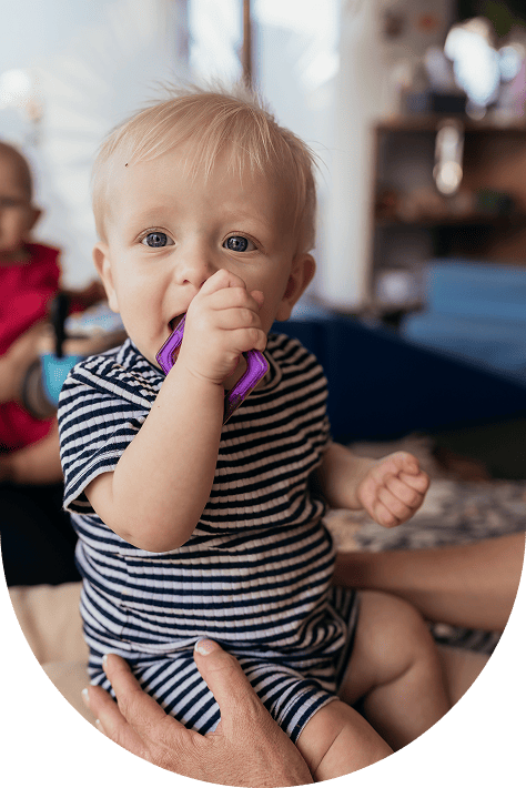Blond toddler in a striped outfit chewing on a purple toy while sitting on an adult's lap indoors.
