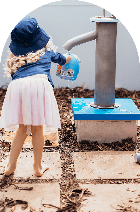 Young girl in a blue hat and pink skirt pouring water from a blue watering can next to a water pump outdoors.