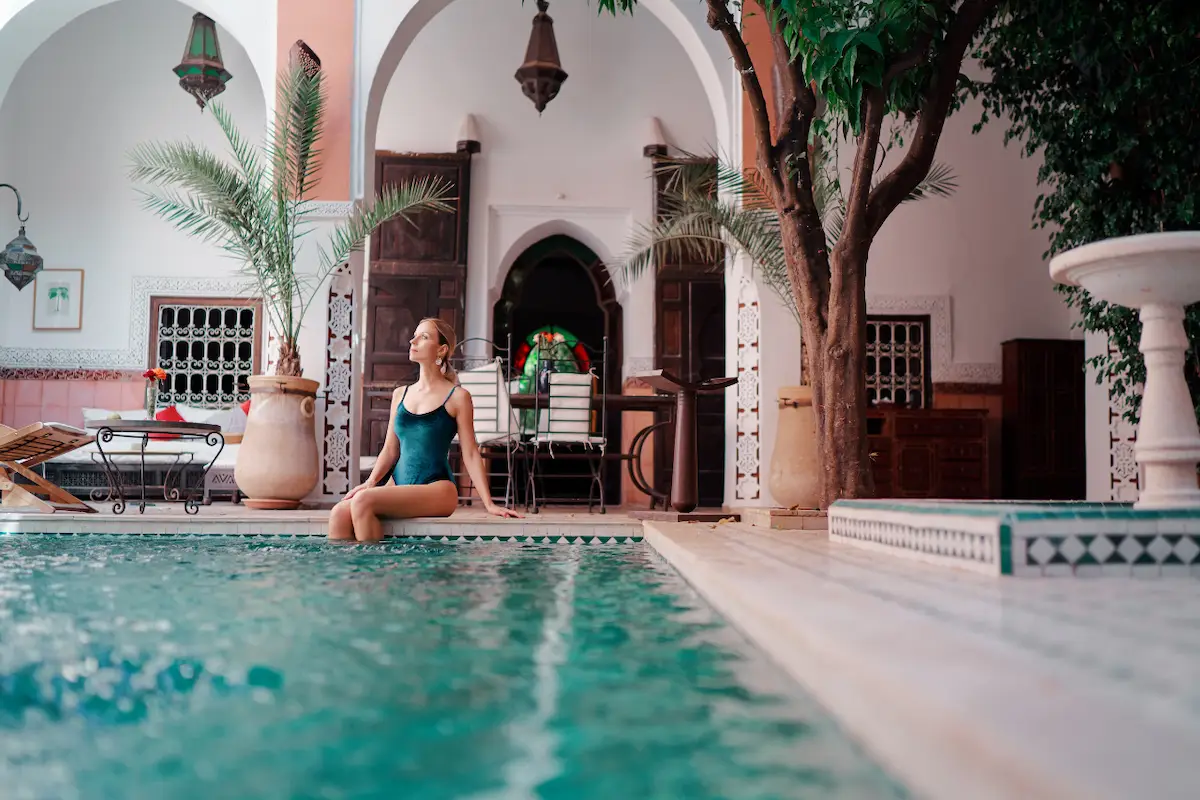 A woman sitting by a swimming pool in a traditional Moroccan-style courtyard riad.