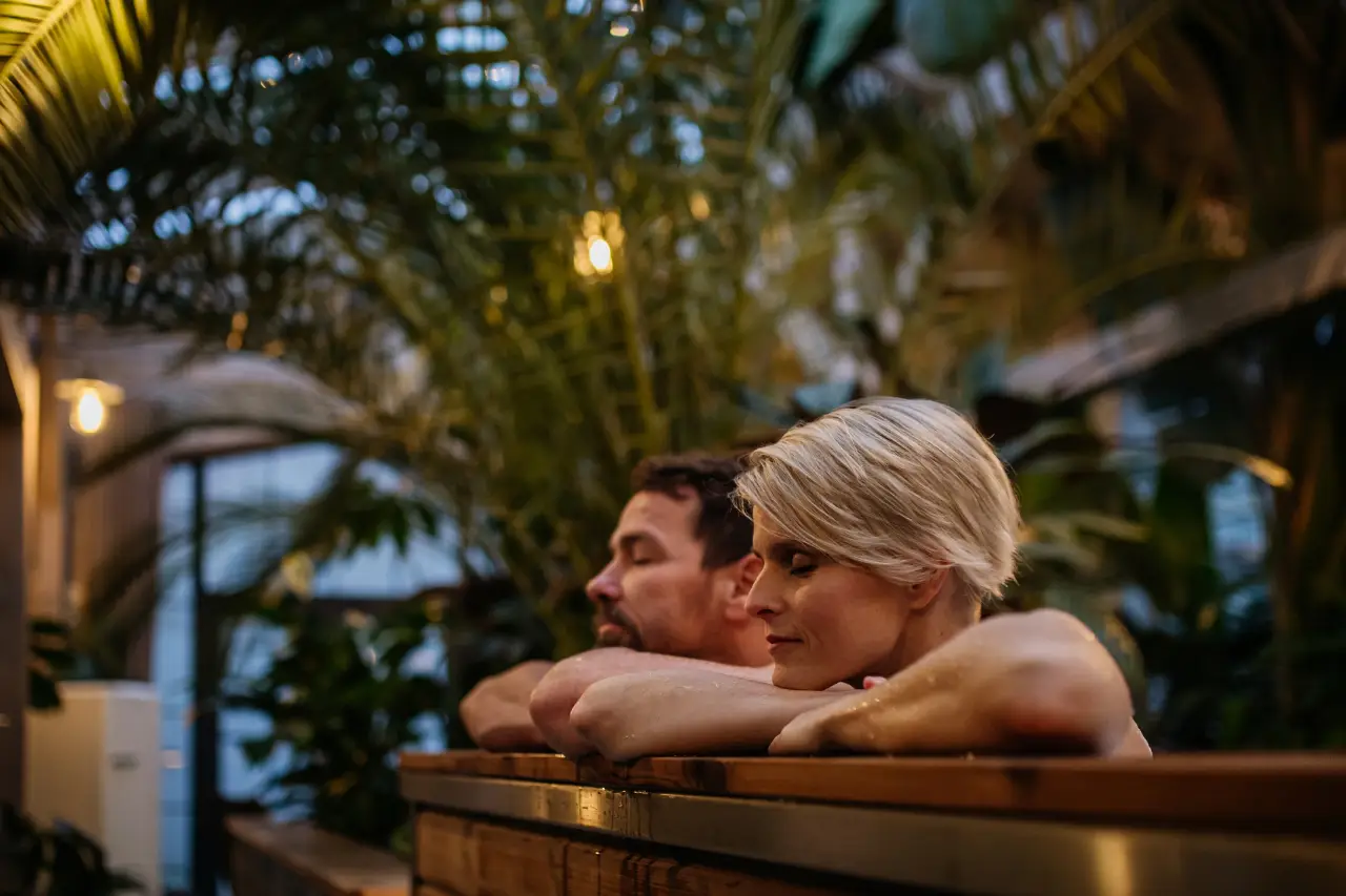 An older couple relaxing in a wooden hot tub surrounded by lush greenery at night.