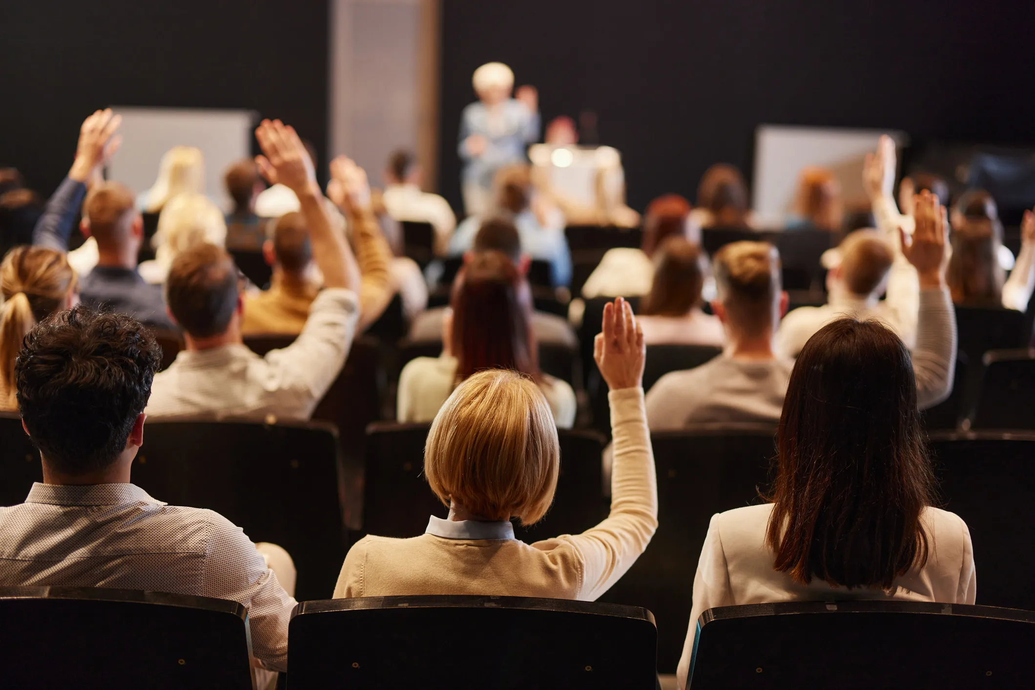Audience raising hands in a conference or seminar room facing a speaker on stage.