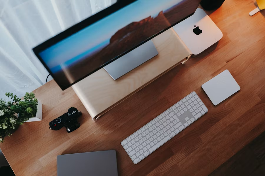 Overhead view of a wooden desk with an Apple monitor, Mac Mini, wireless keyboard, trackpad, laptop, camera, and a small potted plant.