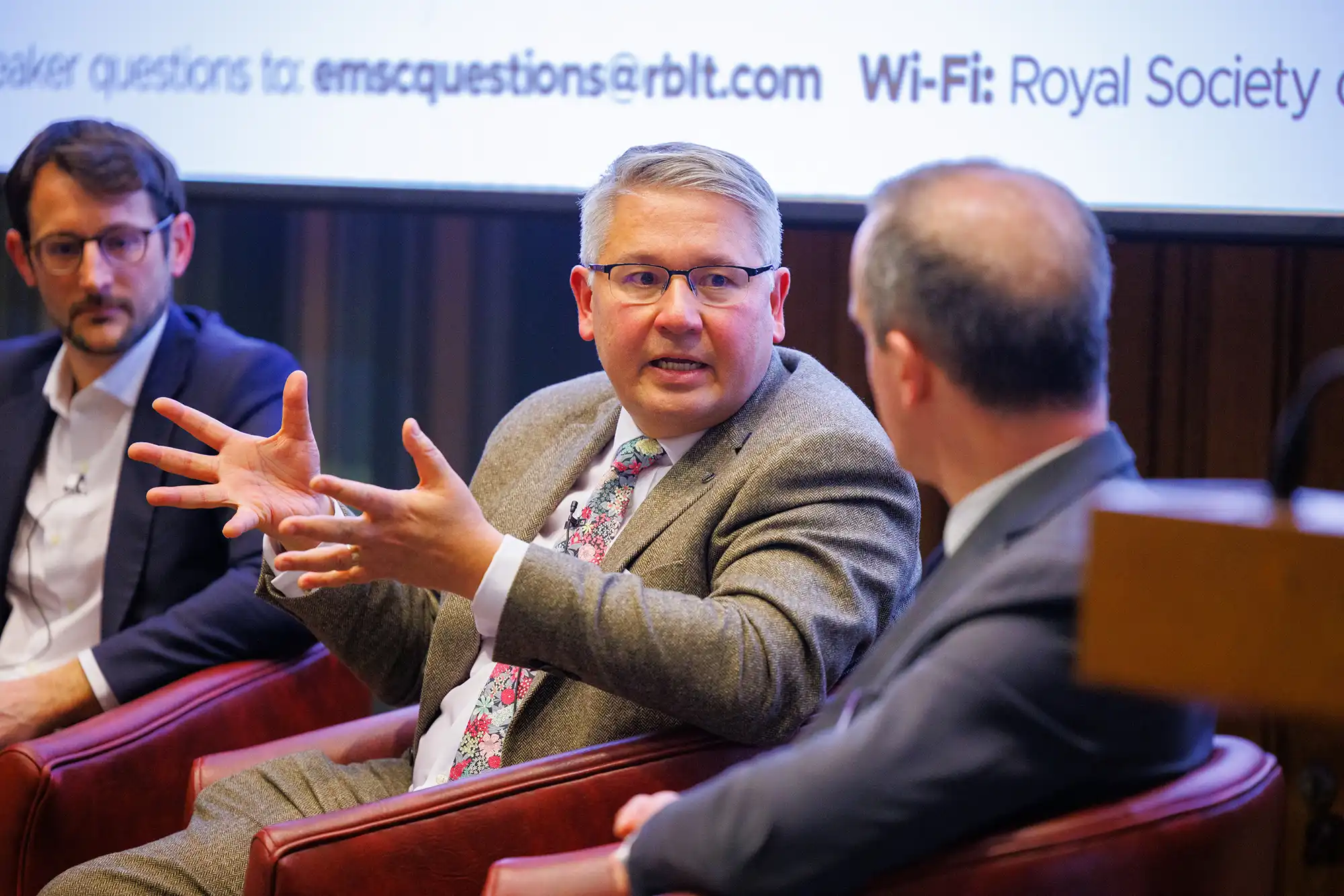 A speaker in a grey blazer and floral tie explains a point with open hands during a panel discussion about European market structure and trading.