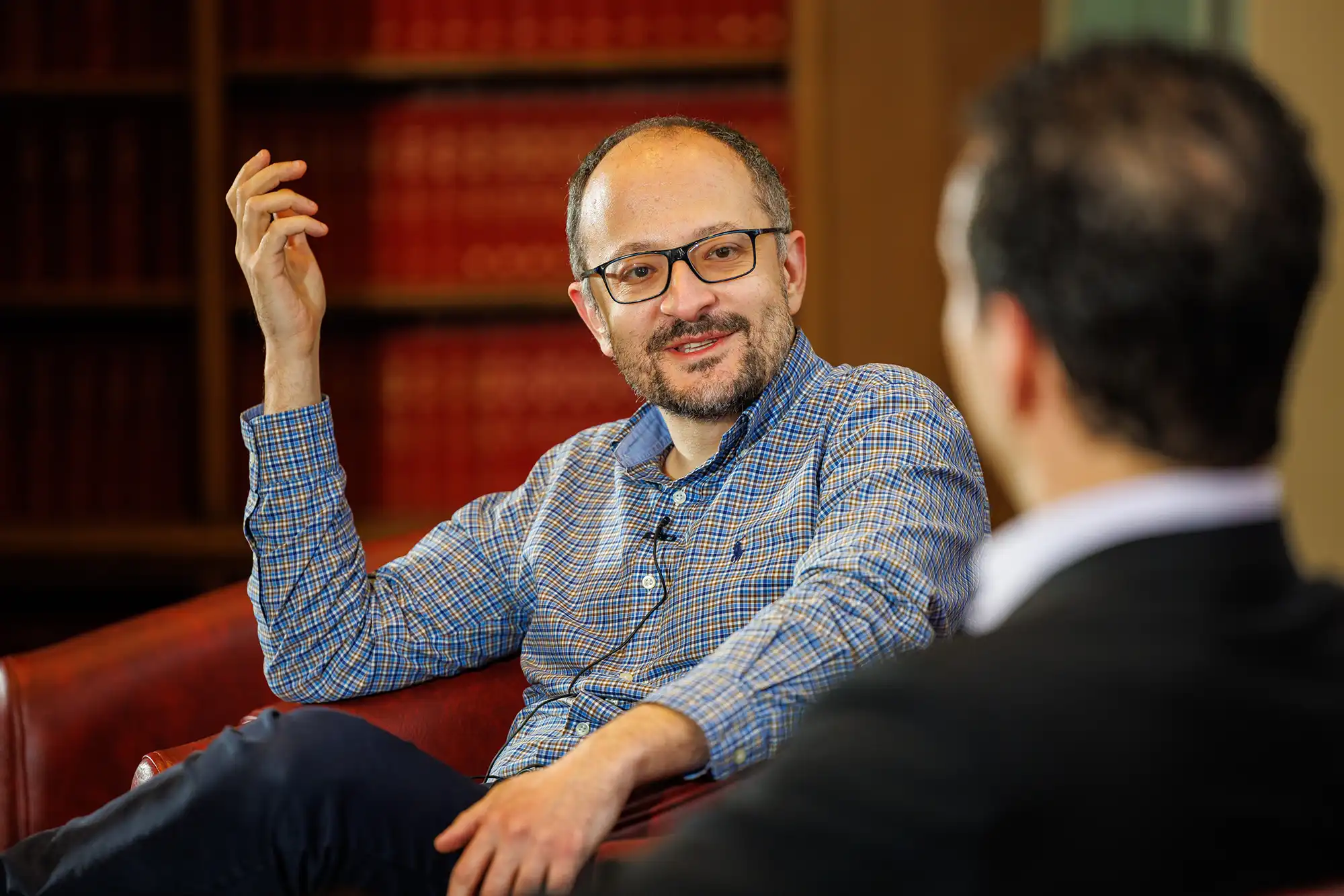 A man in a blue checkered shirt and glasses gestures with one hand while speaking during an informal fireside chat or panel session in a library.