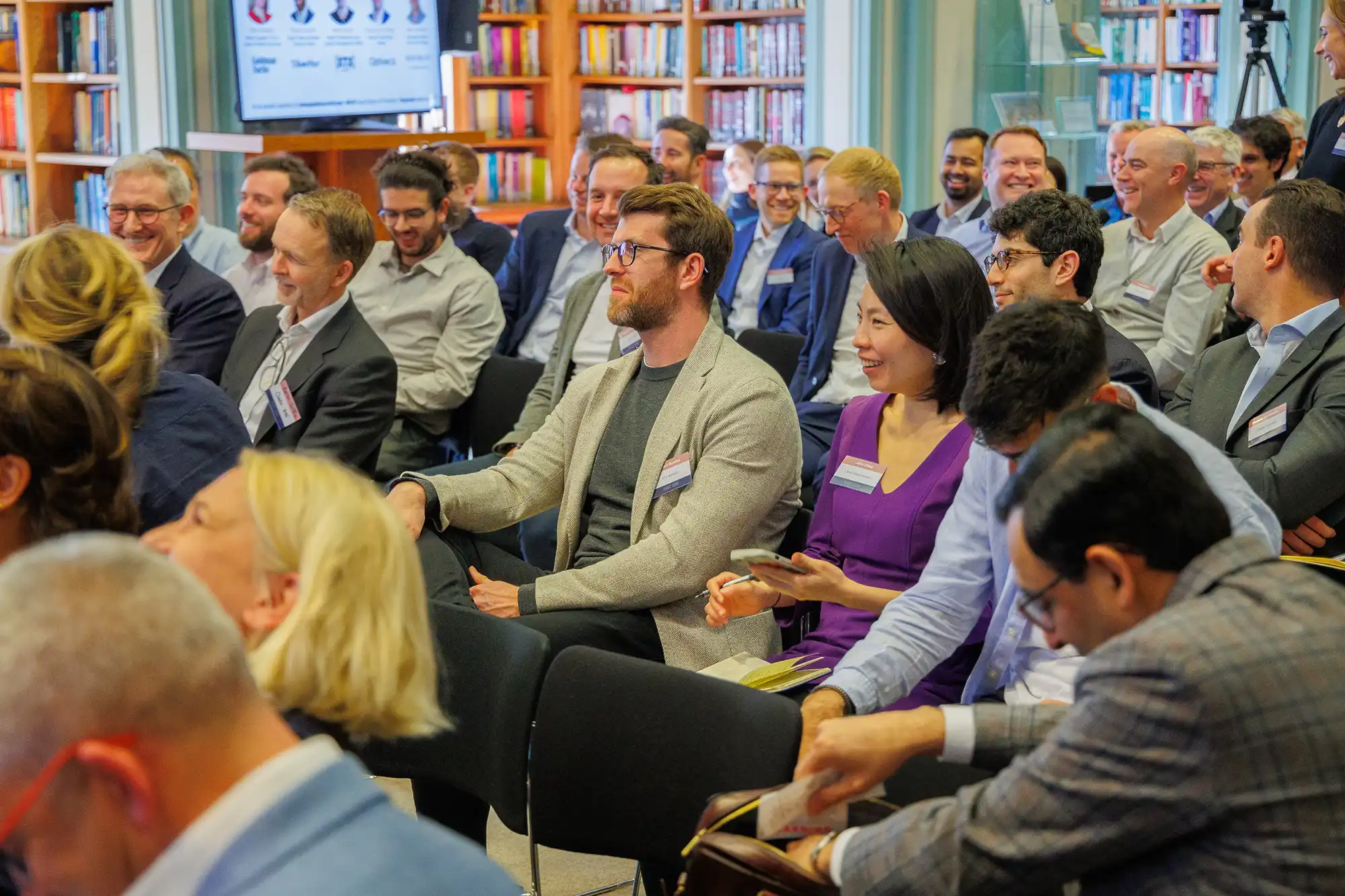 A diverse audience of professionals sits in a library setting, smiling and engaged during a presentation at a European financial trading conference.
