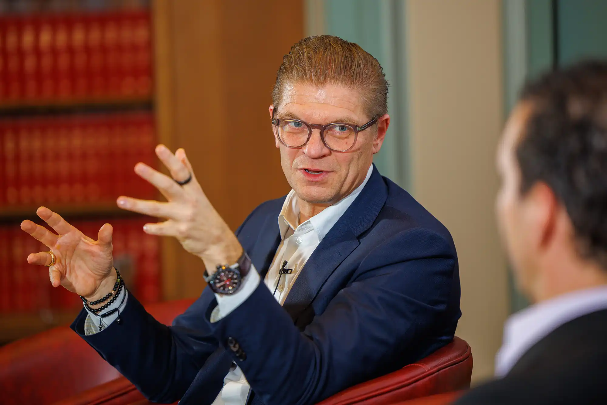 A male speaker with glasses and a blue blazer gestures expressively with both hands while participating in a panel discussion at a library venue.