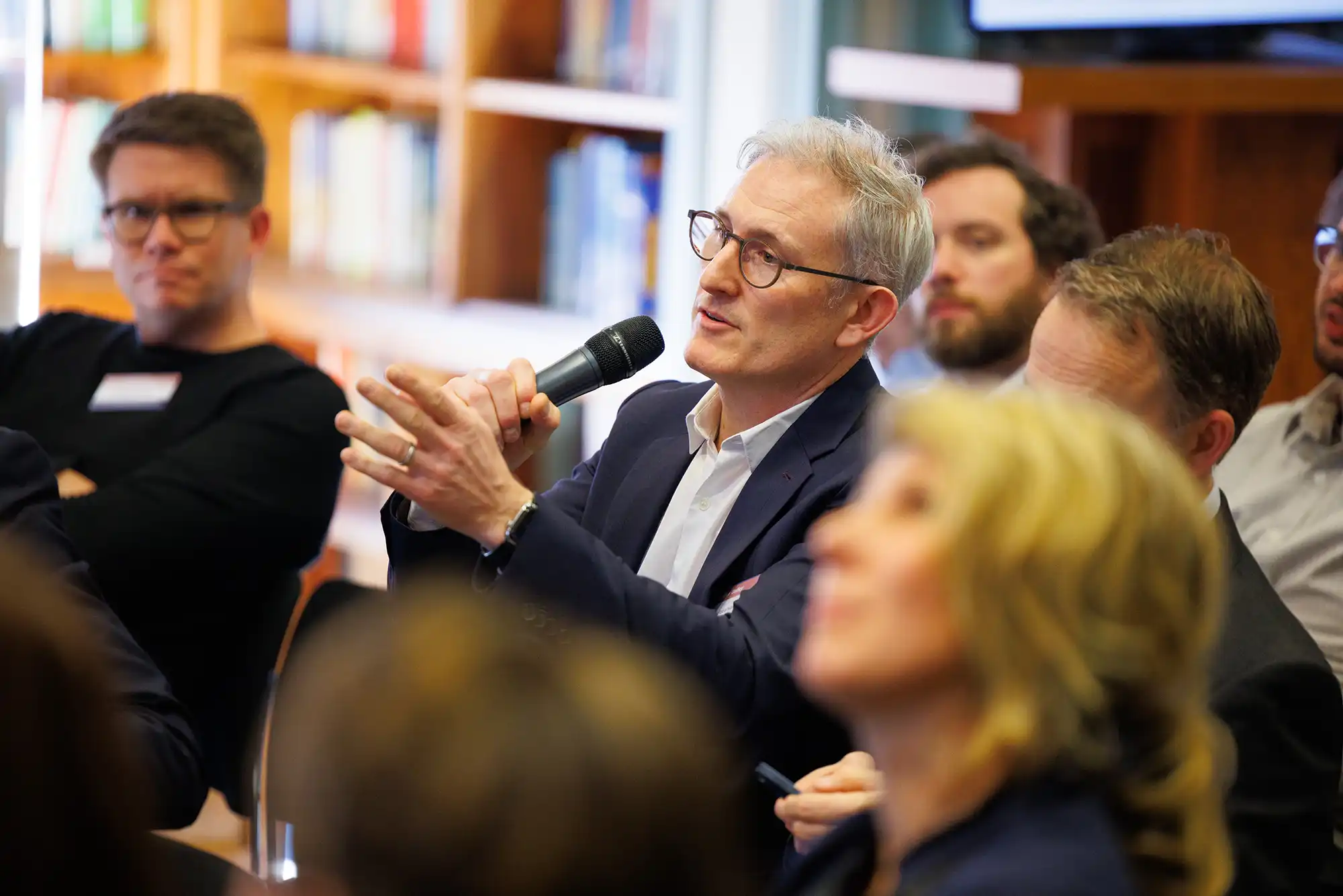 A man with glasses holds a microphone and gestures while asking a question during a Q&A session at a professional financial conference in a library setting.