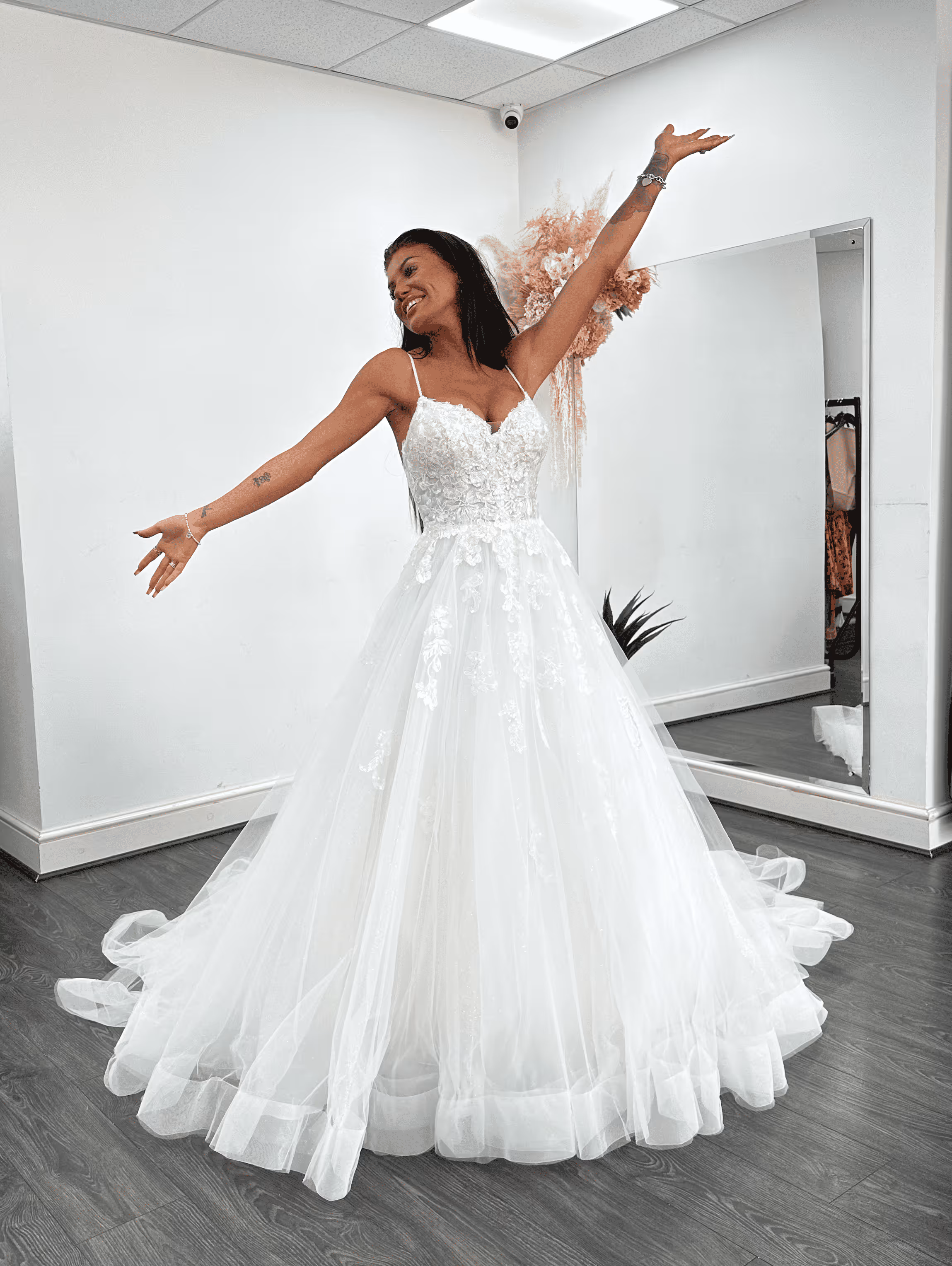 Smiling woman in a white lace wedding gown with arms raised in a fitting room.