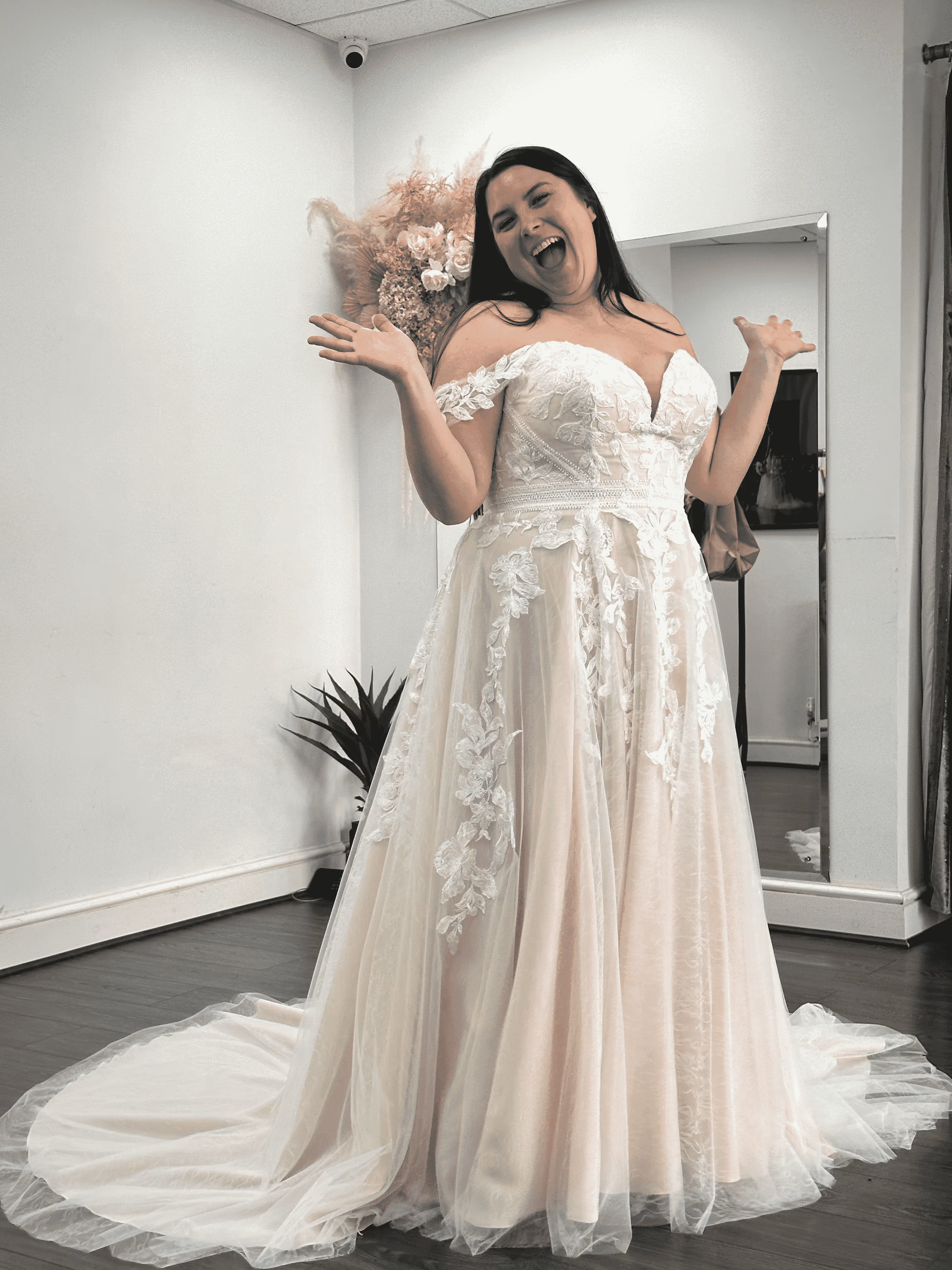 Smiling woman in a white lace off-shoulder wedding dress posing indoors with arms raised.
