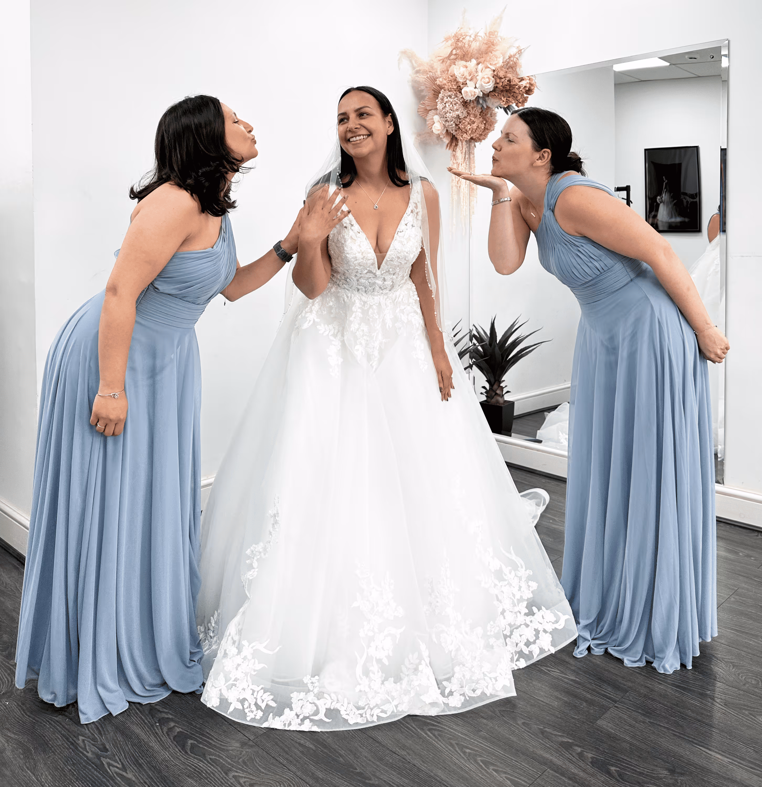 Bride in a white wedding dress smiling as two bridesmaids in blue dresses blow kisses toward her.