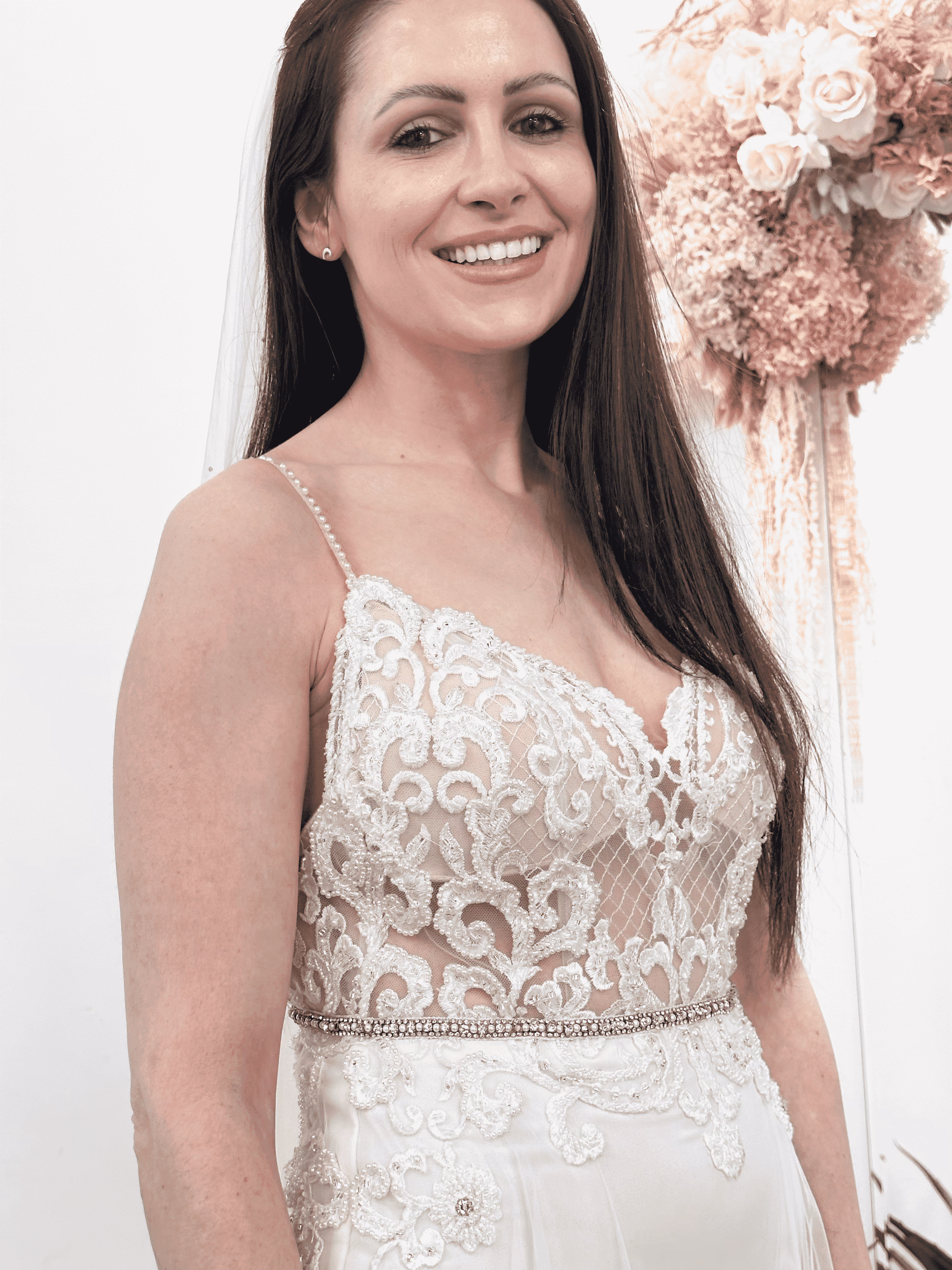 Smiling bride with long dark hair wearing a white lace wedding dress with floral embroidery and thin pearl straps.