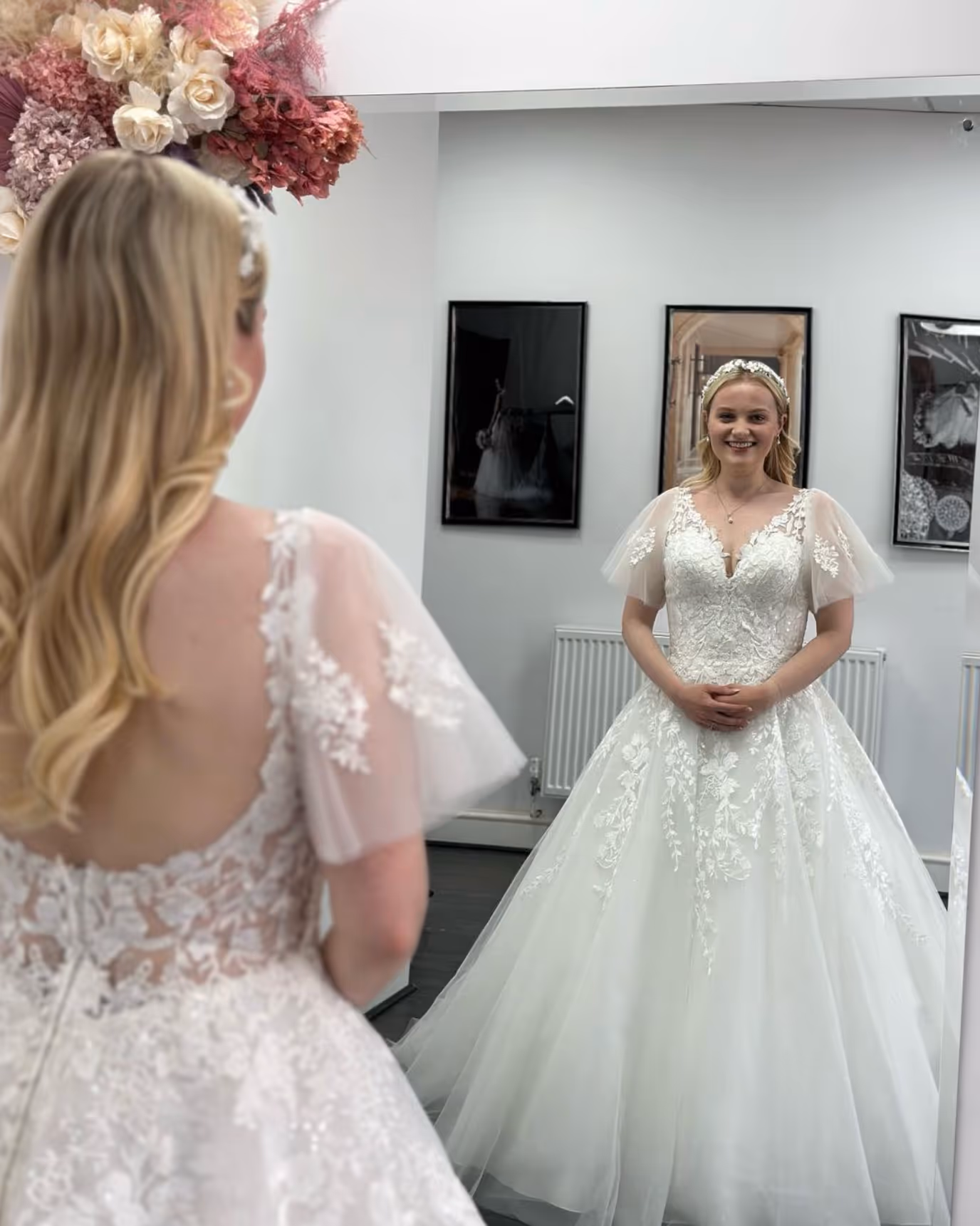 Bride with blonde hair wearing a white lace wedding gown and a jeweled headband, smiling while looking at herself in a mirror inside a room with floral decorations.