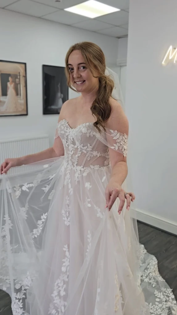 Smiling woman wearing an off-shoulder white wedding dress with floral lace holding the skirt in a bridal shop.