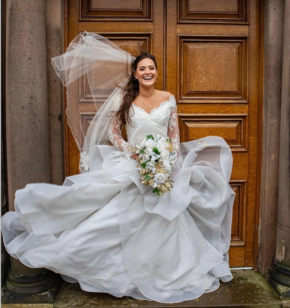 Smiling bride in a flowing white wedding dress and lace sleeves holding a bouquet of white and gold flowers in front of a wooden door.
