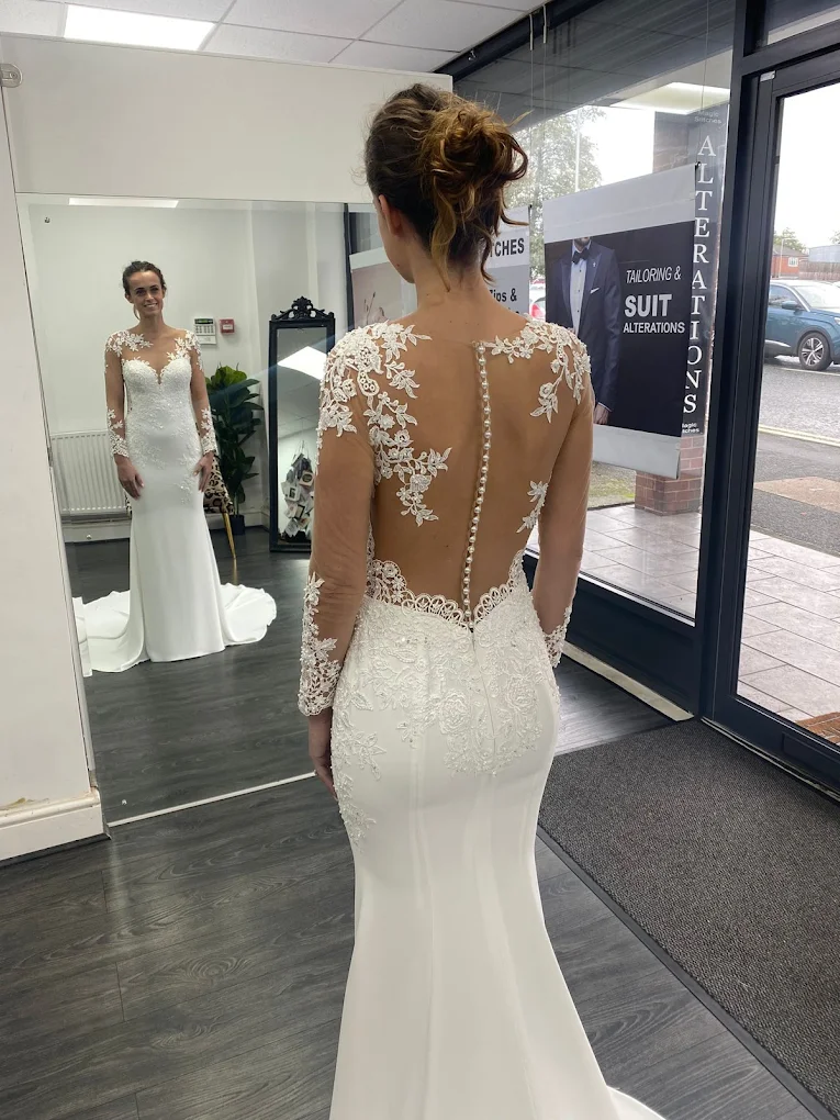 Bride wearing a white lace wedding dress with sheer long sleeves and a buttoned transparent back, standing inside a shop and looking at herself in a mirror.