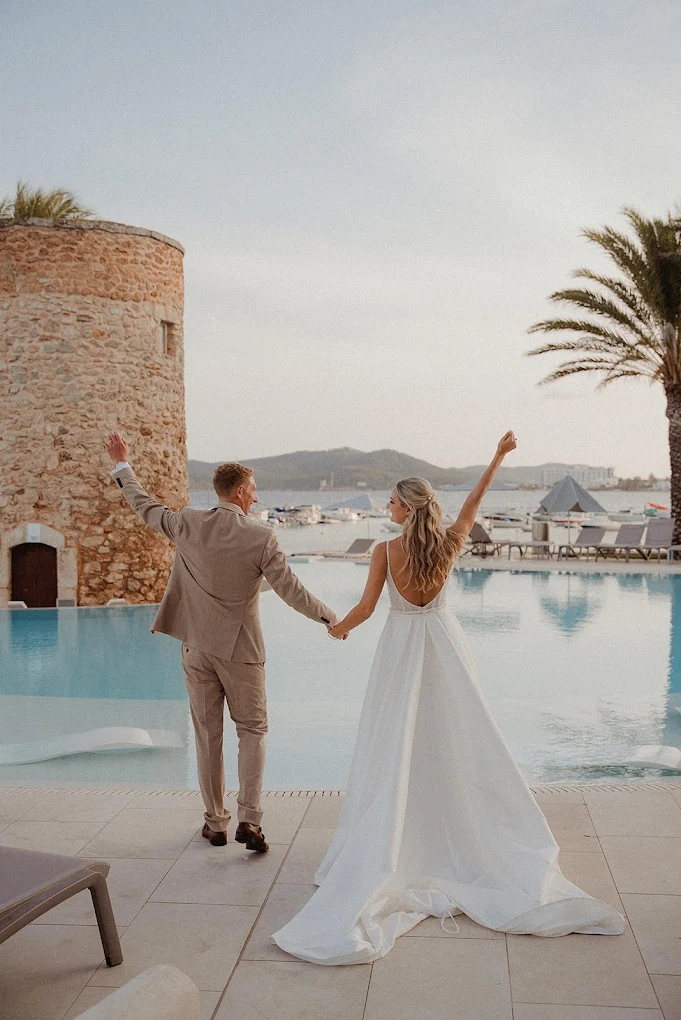 Bride and groom holding hands with raised arms, standing by a pool with a stone tower and palm tree in the background.