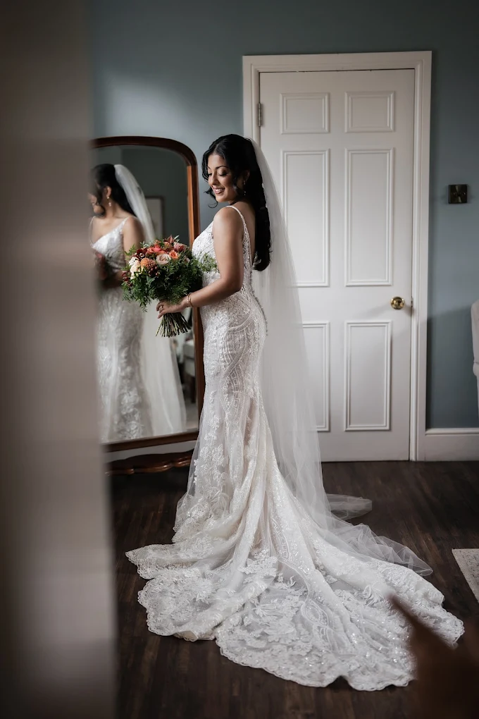 Bride in a white lace wedding gown with a long train and veil holding a bouquet of flowers, standing in front of a mirror.