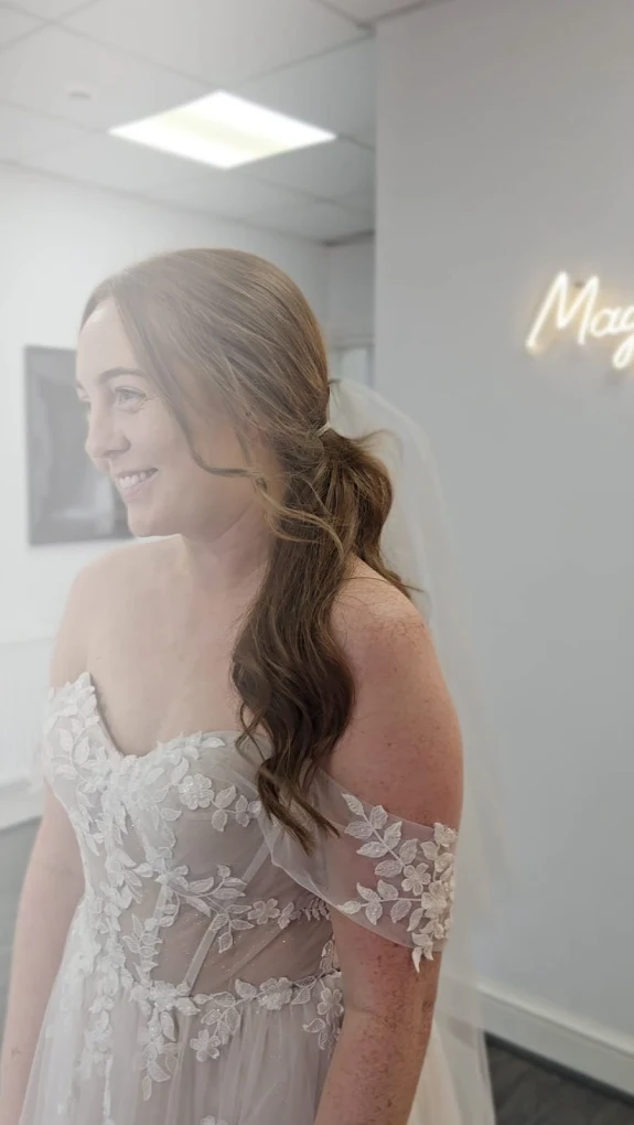 Smiling bride with long wavy hair in an off-shoulder lace wedding dress indoors.