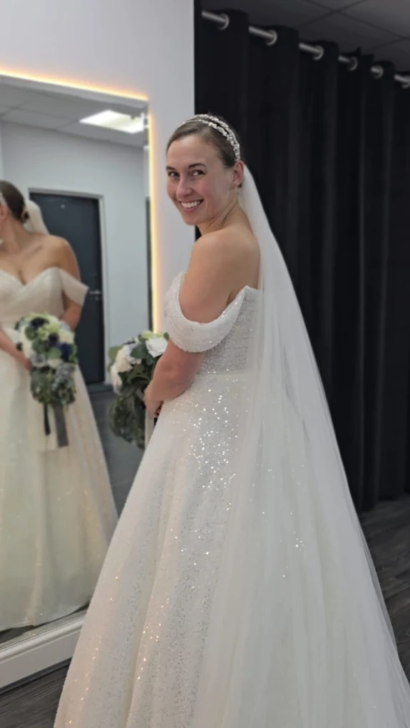 Smiling bride wearing a sparkling off-the-shoulder wedding dress and long veil, standing in front of a mirror holding a bouquet.