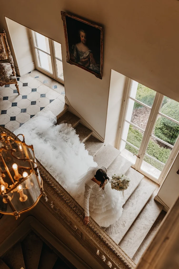 Bride in a white gown holding a bouquet stands on a vintage staircase with large windows and a portrait on the wall.