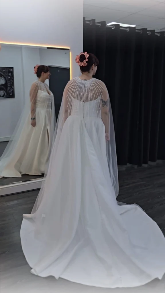Woman wearing a long white wedding dress with a beaded sheer cape, standing in front of a large mirror in a fitting room.