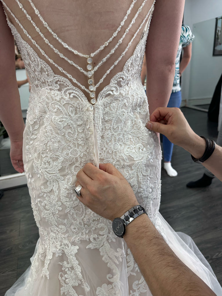 Hands buttoning the back of an ornate lace wedding dress with pearl-like buttons.