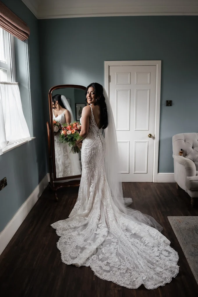 Bride in a lace wedding dress with a long train and veil, holding a bouquet of flowers, standing in front of a mirror in a softly lit room.