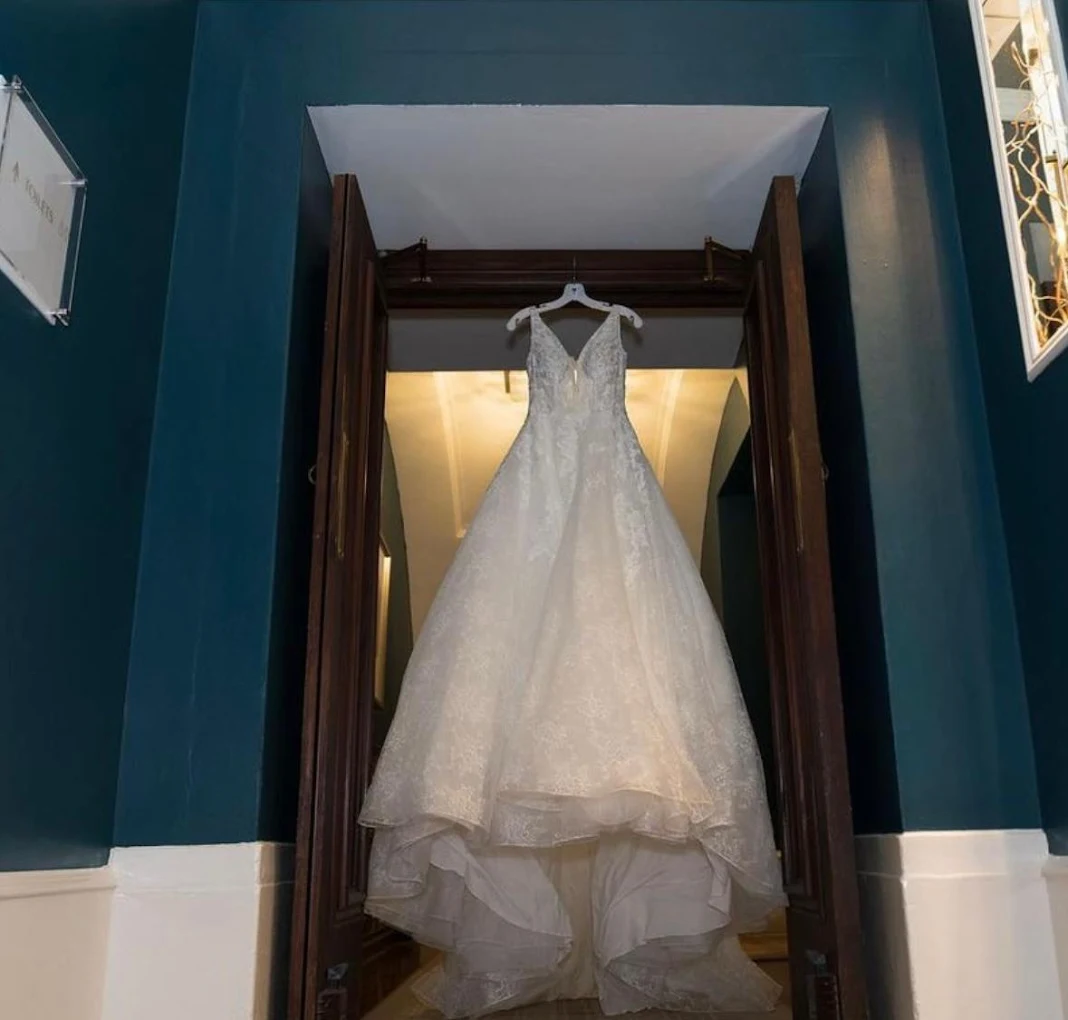 Elegant white lace wedding dress hanging inside a wooden doorway with dark teal walls.