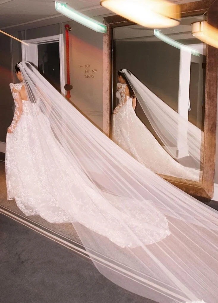 Bride in a lace wedding dress with long train and veil standing in front of a large mirror in a hallway.