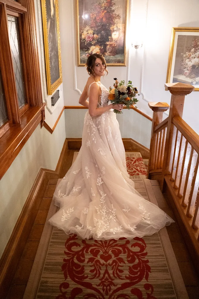 Bride in a white lace wedding dress holding a bouquet, standing on a staircase with wooden railings and framed floral paintings on the walls.