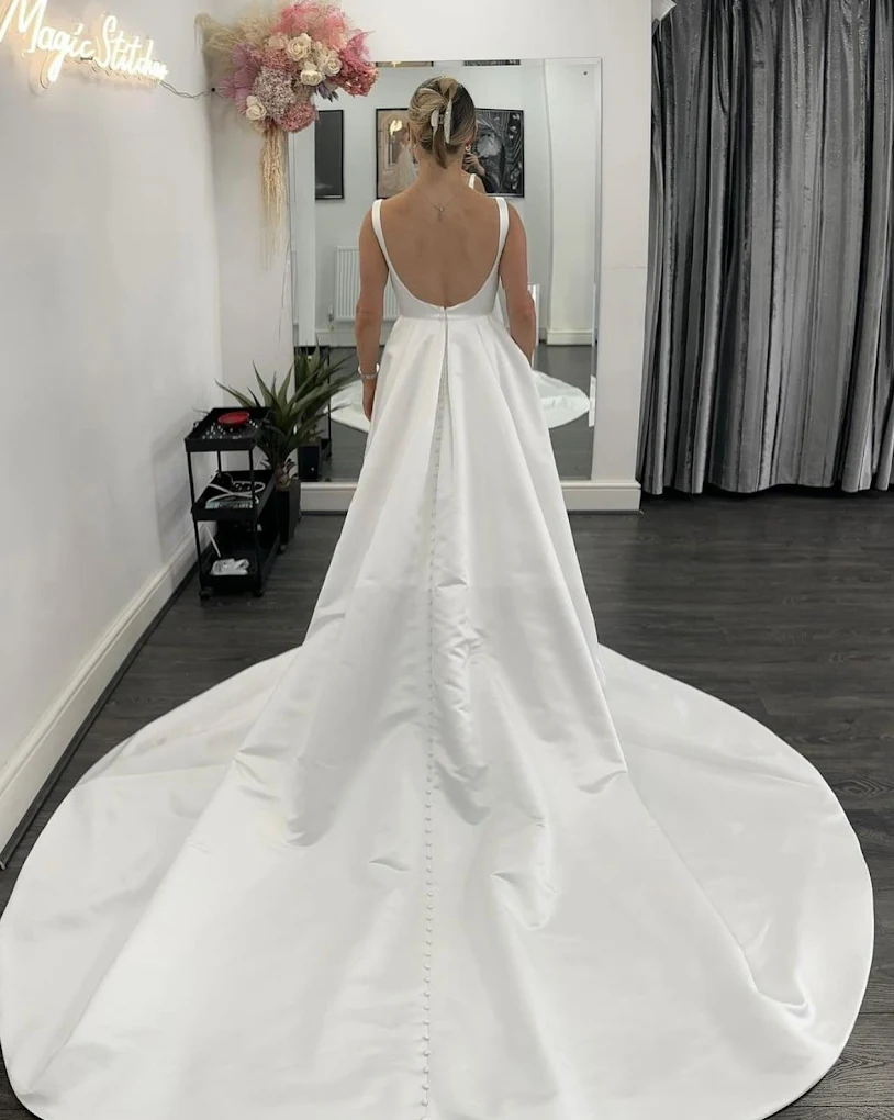 Bride wearing a white satin wedding gown with buttons down the long train, standing facing a mirror in a fitting room.