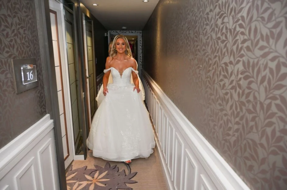 Bride in an off-shoulder white wedding dress walking down a hotel hallway with patterned wallpaper and carpet.