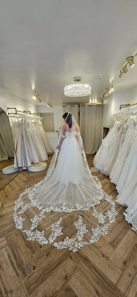 Bride from behind wearing a white wedding gown with a long, lace-embroidered veil, standing in a bridal boutique surrounded by wedding dresses.