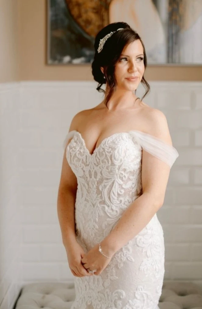 Bride in an off-the-shoulder lace wedding dress with hair styled up and a decorative hairpiece, standing indoors.