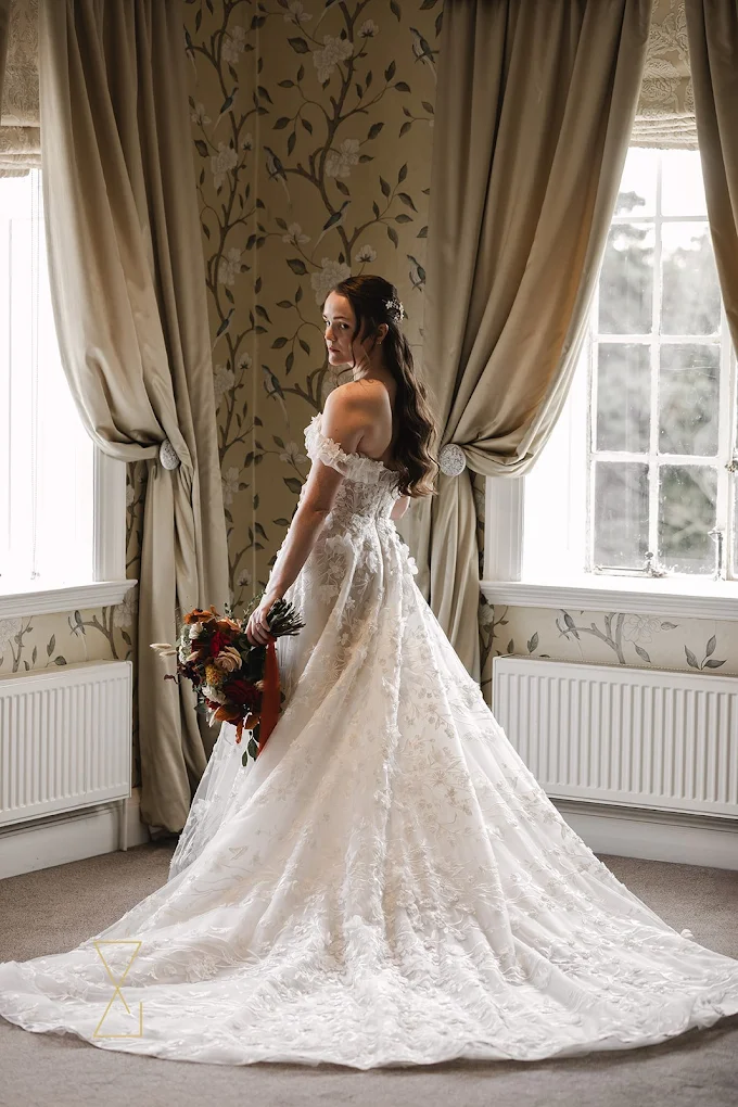 Bride in an off-shoulder white lace wedding gown holding a bouquet, standing between two windows with beige curtains.