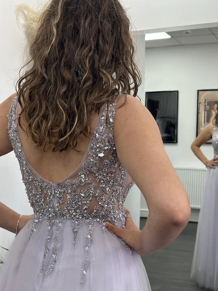 Woman with curly brown hair wearing a sleeveless lavender dress with intricate silver beadwork on the back, standing with hands on hips in front of a mirror.