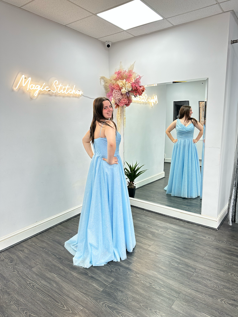 Woman in a sparkling light blue gown posing with hands on hips in front of a large mirror inside a boutique with 'Magic Stitcher' neon sign and floral decor.