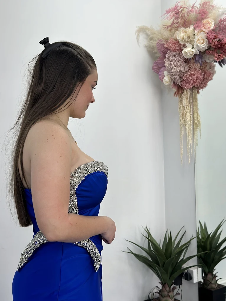 Young woman in a strapless royal blue dress with silver embellishments standing sideways near a white wall with pink and white floral arrangement and two green plants.