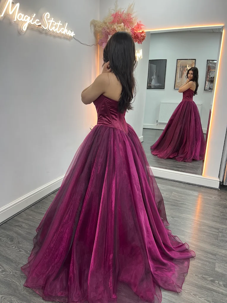 Woman in a strapless magenta ball gown looking at herself in a large mirror in a boutique with a 'Magic Stitches' neon sign on the wall.