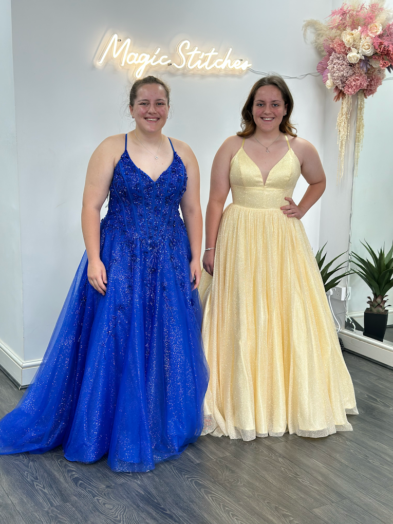 Two young women smiling, wearing floor-length glittery ball gowns in blue and yellow, standing under a neon sign that reads 'Magic Stitches'.