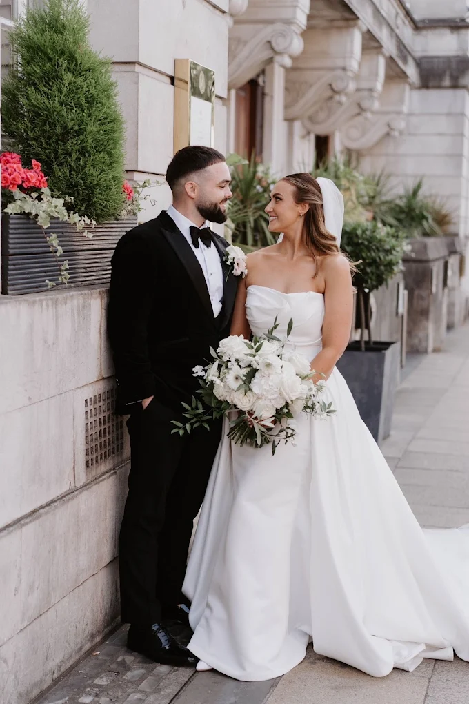 Bride in white strapless gown holding a bouquet and groom in black tuxedo looking at each other outside a stone building.