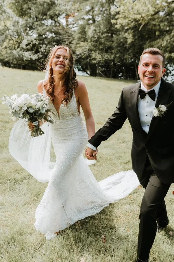Smiling bride in lace wedding dress holding bouquet and groom in black tuxedo holding hands outdoors on grass.