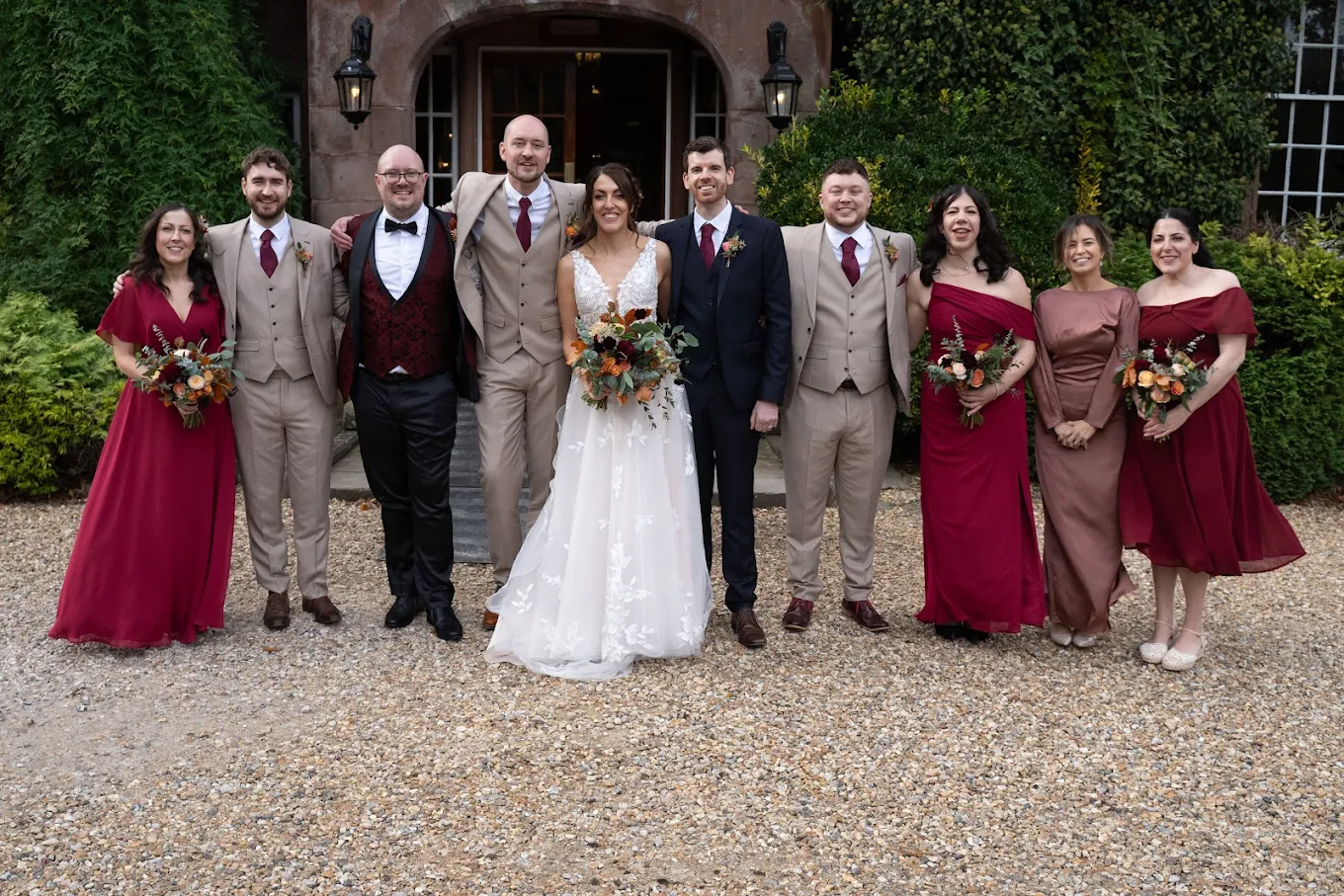 Bride and groom with wedding party dressed in beige suits and burgundy dresses standing outdoors in front of a stone building entrance.