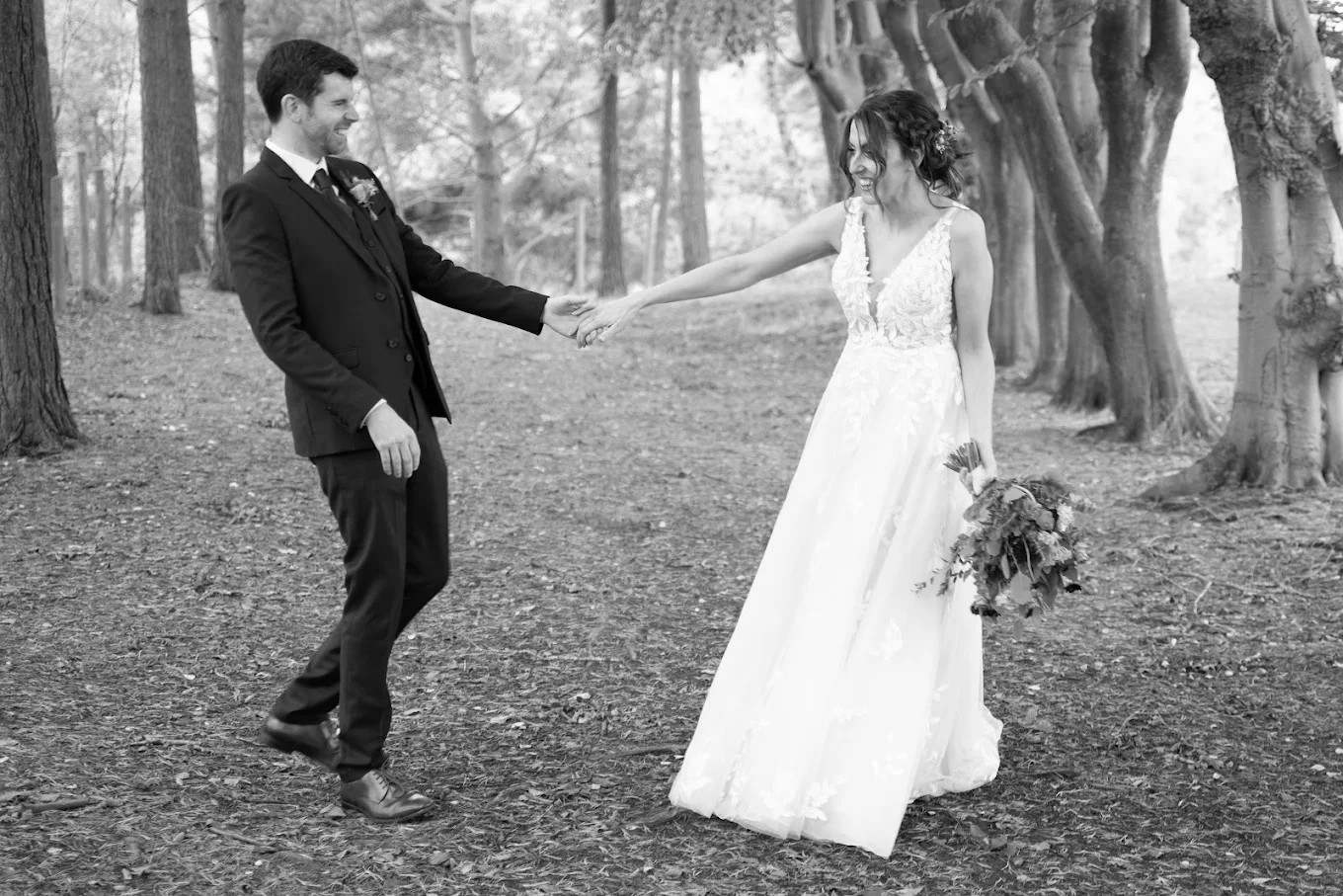 Bride in a white dress holding a bouquet and hand of groom in a suit, standing on a wooded path.